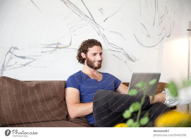 Lächelnder junger Mann sitzt mit Laptop auf der Couch in einem Café Cafe Kaffeehaus Bistro Cafes Kaffeehäuser Notebook Laptops Notebooks Männer männlich