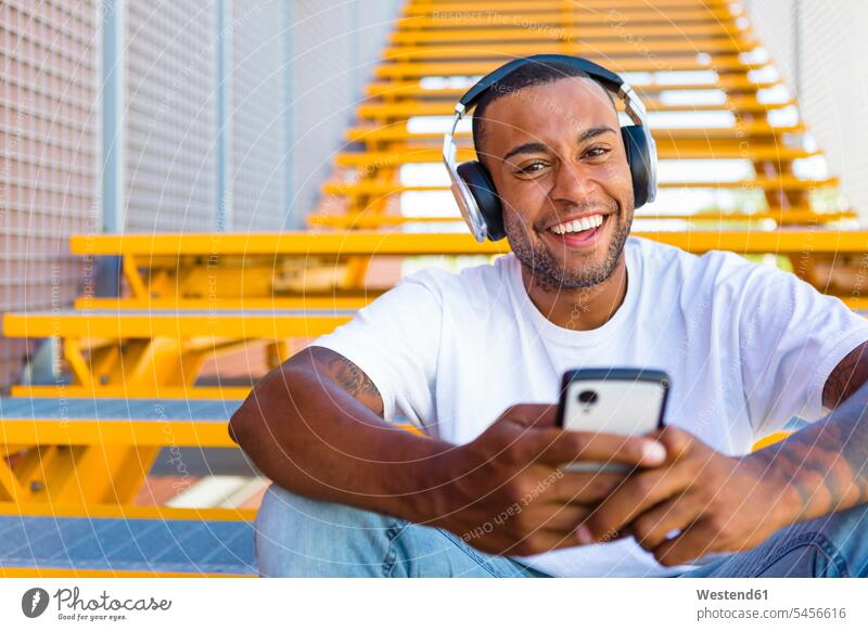 Porträt eines lachenden jungen Mannes mit Kopfhörer und Smartphone, der auf einer Treppe sitzt iPhone Smartphones Portrait Porträts Portraits Männer männlich