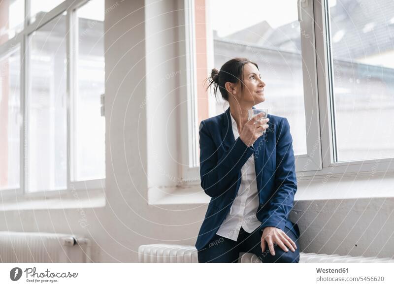 Geschäftsfrau mit einem Glas Wasser, die auf einer Heizung auf einem Dachboden sitzt und durch ein Fenster schaut Geschäftsfrauen Businesswomen Businessfrauen
