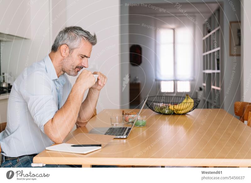 Lächelnder Mann trinkt Kaffee und benutzt Laptop im Heimbüro lächeln trinken Home Office homeoffice zuhause zuhause arbeiten arbeiten von zuhause Zuhause