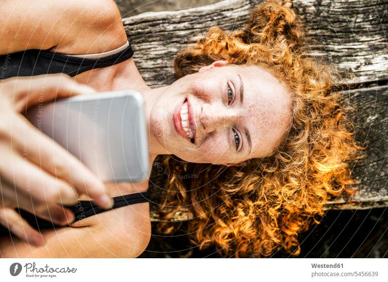 Porträt einer rothaarigen jungen Frau, die mit ihrem Handy auf einer Bank liegt Portrait Porträts Portraits weiblich Frauen Erwachsener erwachsen Mensch