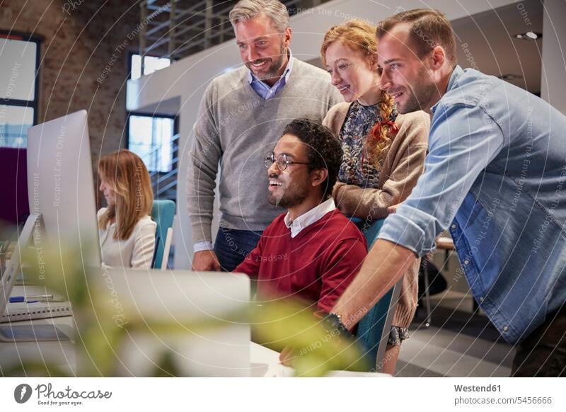 Kollegen schauen einem jungen Mann über die Schulter, der in einem modernen Büro arbeitet PC Personal Computers Desktop PC Kollegin Kolleginnen lachen Team