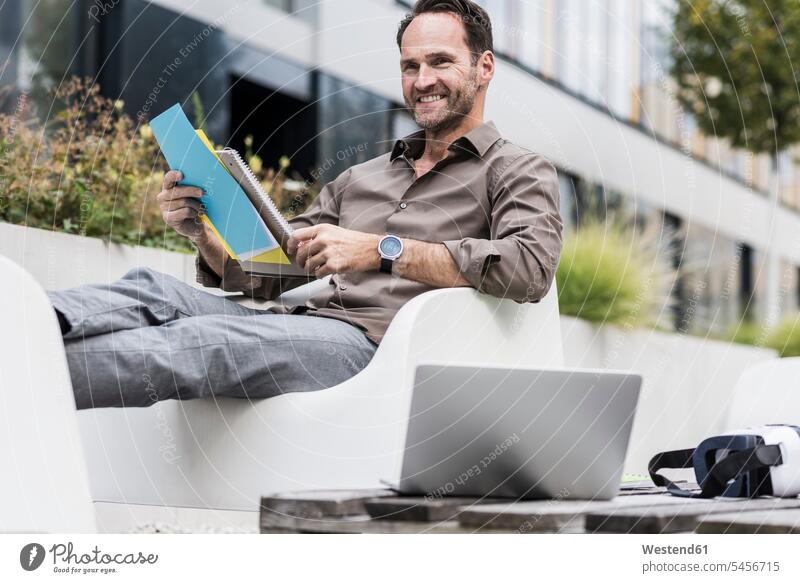 Lächelnder Geschäftsmann mit Dokument, Laptop und Virtual-Reality-Brille auf der Terrasse sitzend Businessmann Businessmänner Geschäftsmänner Geschäftsleute