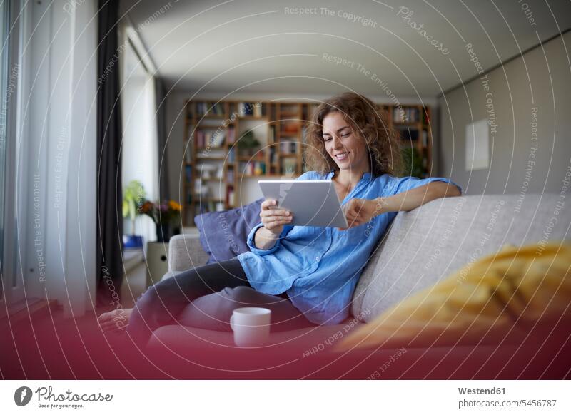 Frau benutzt digitales Tablet, während sie zu Hause auf dem Sofa sitzt Farbaufnahme Farbe Farbfoto Farbphoto Innenaufnahme Innenaufnahmen innen drinnen Tag