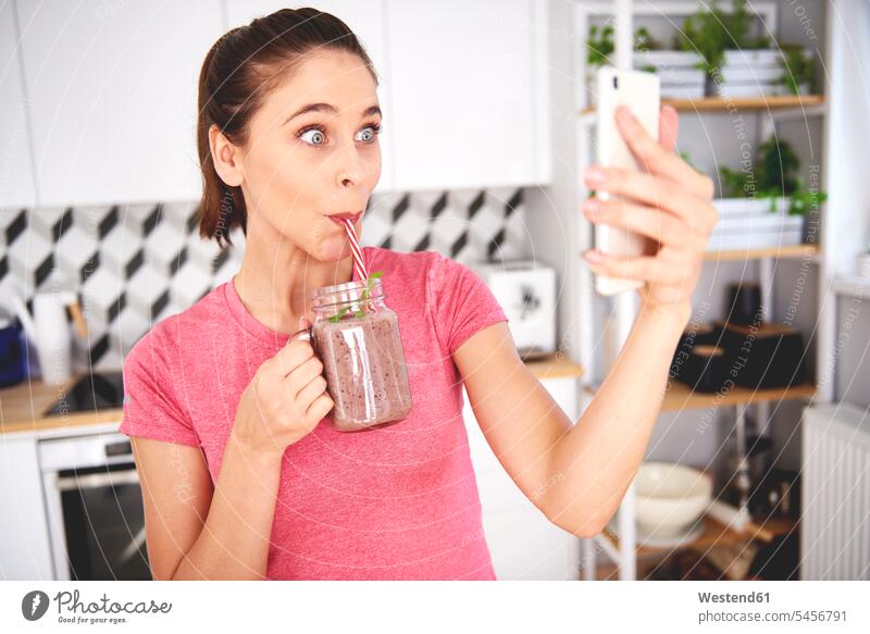 Porträt einer jungen Frau, die ein Selfie mit ihrem Smartphone in der Küche macht, während sie einen Smoothie trinkt trinken Küchen Portrait Porträts Portraits