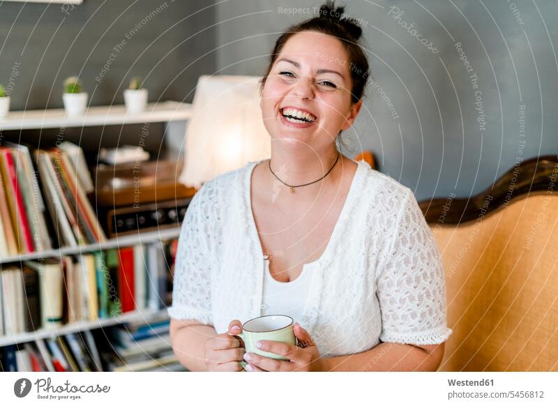 Fröhliche, sinnliche junge Frau hält Kaffeetasse, während sie im Café sitzt Farbaufnahme Farbe Farbfoto Farbphoto Innenaufnahme Innenaufnahmen innen drinnen