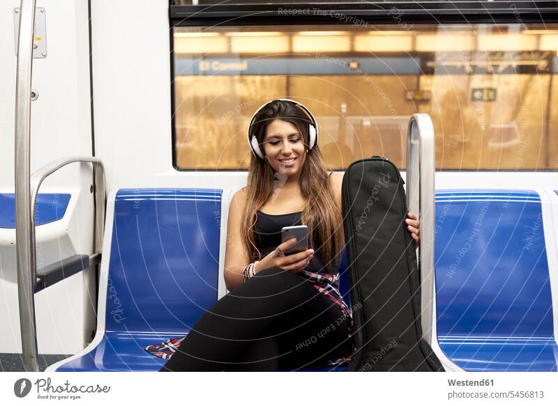 Porträt einer lächelnden Frau mit Gitarre und Kopfhörer, die in der U-Bahn auf ein Handy schaut weiblich Frauen Portrait Porträts Portraits ansehen Smartphone