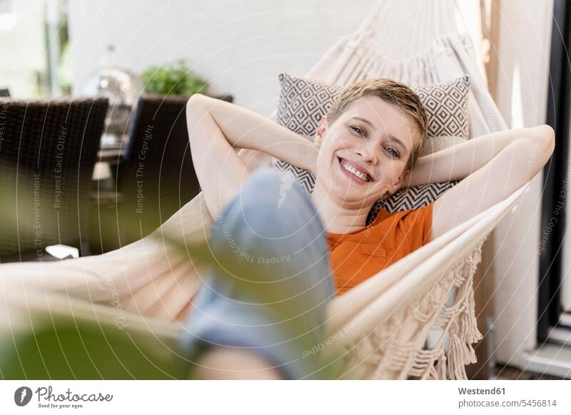 Lächelnde, mittelgroße, erwachsene Frau mit Händen hinter dem Kopf auf der Hängematte in der Veranda Farbaufnahme Farbe Farbfoto Farbphoto Deutschland