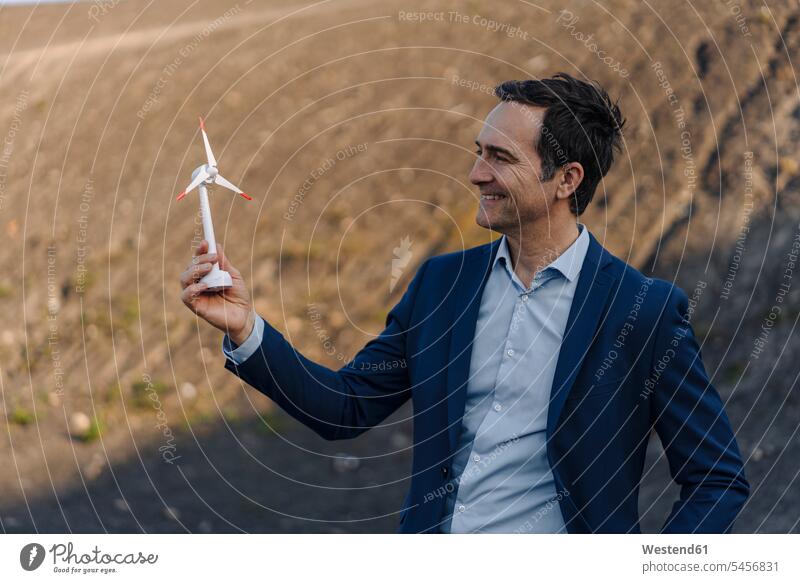 Glücklicher reifer Geschäftsmann auf einer stillgelegten Minenhalde mit einem Modell einer Windkraftanlage Leute Menschen People Person Personen Europäisch