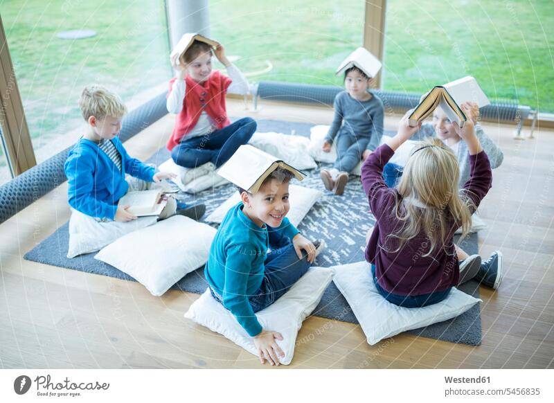 Lächelnde Schülerinnen und Schüler sitzen im Pausenraum der Schule auf dem Boden und balancieren Bücher auf ihren Köpfen Böden Boeden lächeln sitzend sitzt