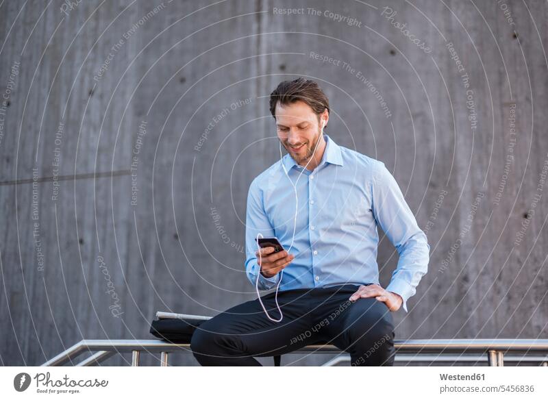 Lächelnder Geschäftsmann mit Ohrstöpseln, der auf einem Geländer sitzt und auf sein Smartphone schaut Businessmann Businessmänner Geschäftsmänner iPhone