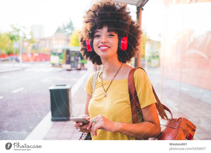 Glückliche junge Frau mit Afrofrisur, die in der Stadt mit Kopfhörern Musik hört Leute Menschen People Person Personen gelockt gelockte Haare gelocktes Haar