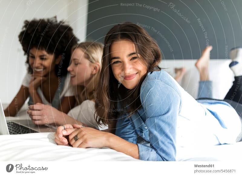 Porträt einer lächelnden jungen Frau, die mit Freundinnen auf dem Bett liegt und einen Laptop benutzt weiblich Frauen Notebook Laptops Notebooks liegen liegend