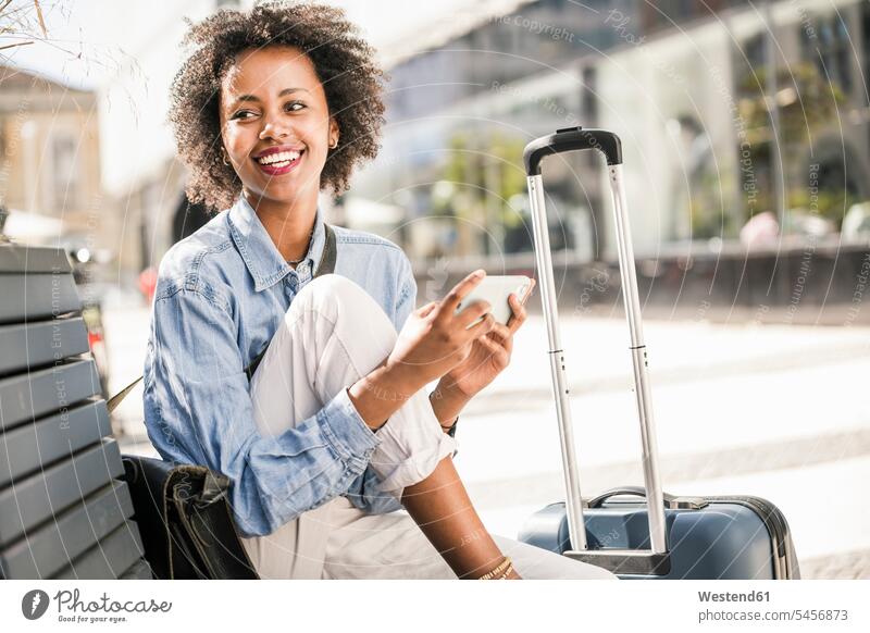 Glückliche junge Frau sitzt auf einer Bank mit Handy und Koffer Leute Menschen People Person Personen Afrikanisch Afrikanische Abstammung dunkelhäutig Farbige
