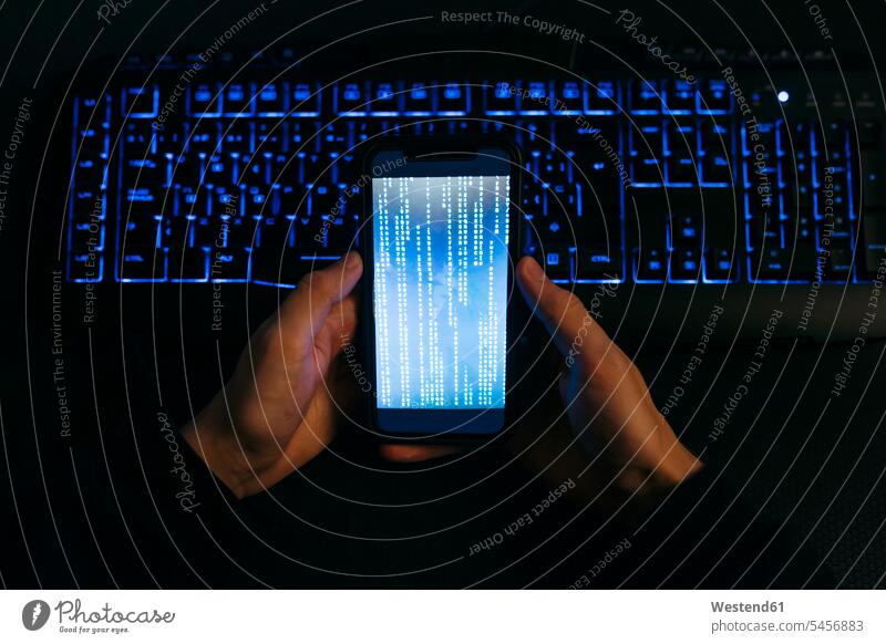 Computerhacker hält Mobiltelefon mit der Hand gegen Computertastatur im Büro Farbaufnahme Farbe Farbfoto Farbphoto Innenaufnahme Innenaufnahmen innen drinnen