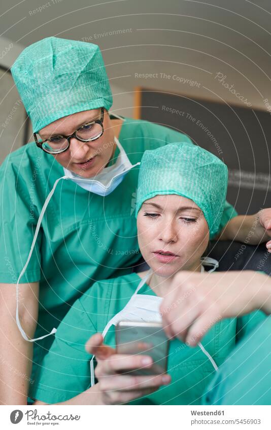 Zwei Frauen in Kitteln schauen auf ein Mobiltelefon Gesundheit Deutschland benutzen Beruf Berufstätigkeit Berufe Beschäftigung Jobs Schutz geschützt schützen
