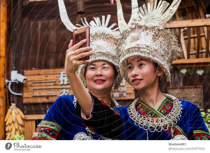 China, Guizhou, zwei Miao-Frauen in traditionellen Kleidern und mit Kopfschmuck machen ein Selfie mit Smartphone Einheimische einheimisch Einheimischer