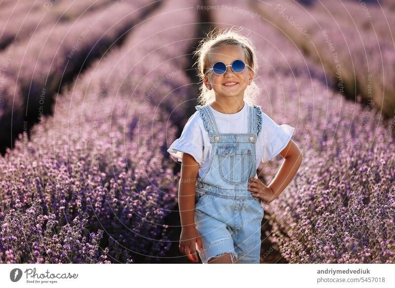 adorable Kind Mädchen in Lavendelfeld auf Sonnenuntergang. lächelnd Kind in Sonnenbrille, Jeans Overall hat Spaß an der Natur am Sommertag. Familientag, Urlaub, holiday.International Children's Day,