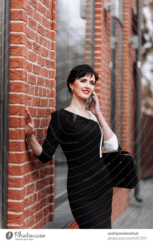 Glücklicher Anwalt Geschäftsfrau professionell ist zu Fuß im Freien im Gespräch auf Zelle Smartphone. Kaukasische erfolgreiche Frau lächelnd trägt stilvolle weiß-schwarzes Kleid mit schwarzer Tasche