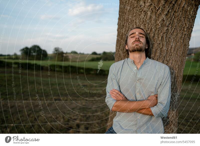 Entspannter junger Mann lehnt an einem Baumstamm auf dem Land Leute Menschen People Person Personen Europäisch Kaukasier kaukasisch 1 Ein ein Mensch