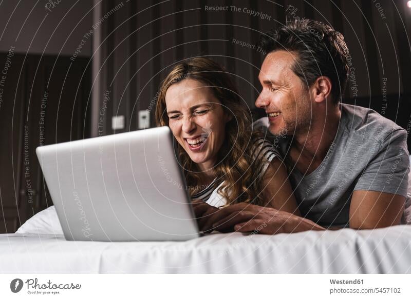 Glückliches Paar liegt zu Hause auf dem Bett und hat Spaß mit dem Laptop Zuhause daheim Notebook Laptops Notebooks liegen liegend Betten Spass Späße spassig