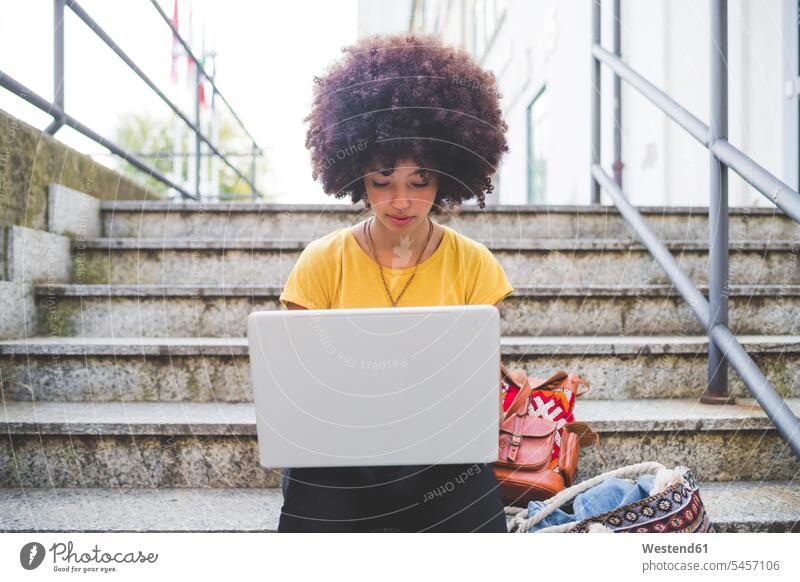 Junge Frau mit Afrofrisur mit Laptop auf einer Treppe in der Stadt Leute Menschen People Person Personen gelockt gelockte Haare gelocktes Haar lockig