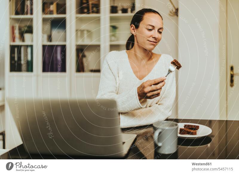 Frau isst Schokoladen-Brownie, während sie mit Kaffee und Laptop in beleuchteter Küche sitzt Brownies Süßigkeiten Süßes Essen Food Food and Drink