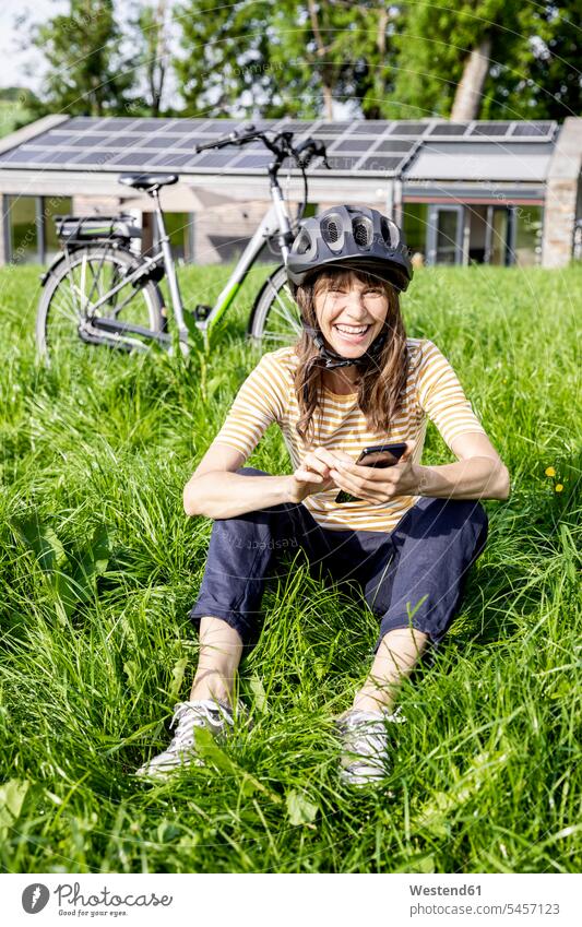 Lachende Frau mit Fahrrad mit Handy auf einer Wiese vor einem Haus Leute Menschen People Person Personen Europäisch Kaukasier kaukasisch 1 Ein ein Mensch