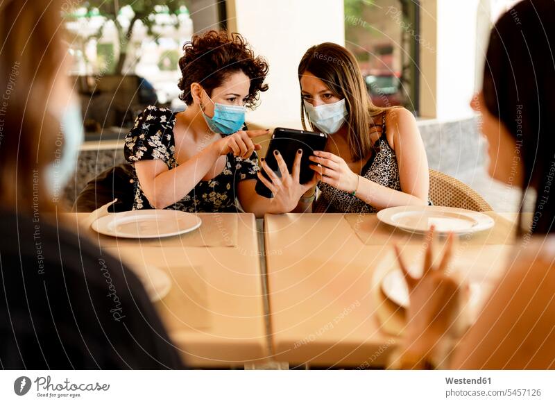 Frauen, die Masken tragen, während sie am digitalen Tablet mit Freunden am Tisch im Restaurant sitzen Farbaufnahme Farbe Farbfoto Farbphoto Spanien