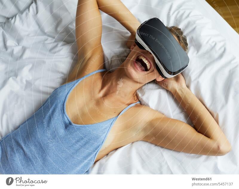 Lachende junge Frau mit VR-Brille im Bett liegend Brillen lachen weiblich Frauen Betten glücklich Glück glücklich sein glücklichsein liegt positiv Emotion