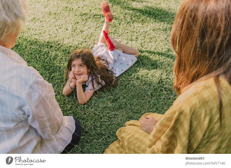 Hübsches Mädchen mit Händen am Kinn, das sich mit der Familie unterhält, während es über Grasland im Hof liegt Farbaufnahme Farbe Farbfoto Farbphoto Spanien