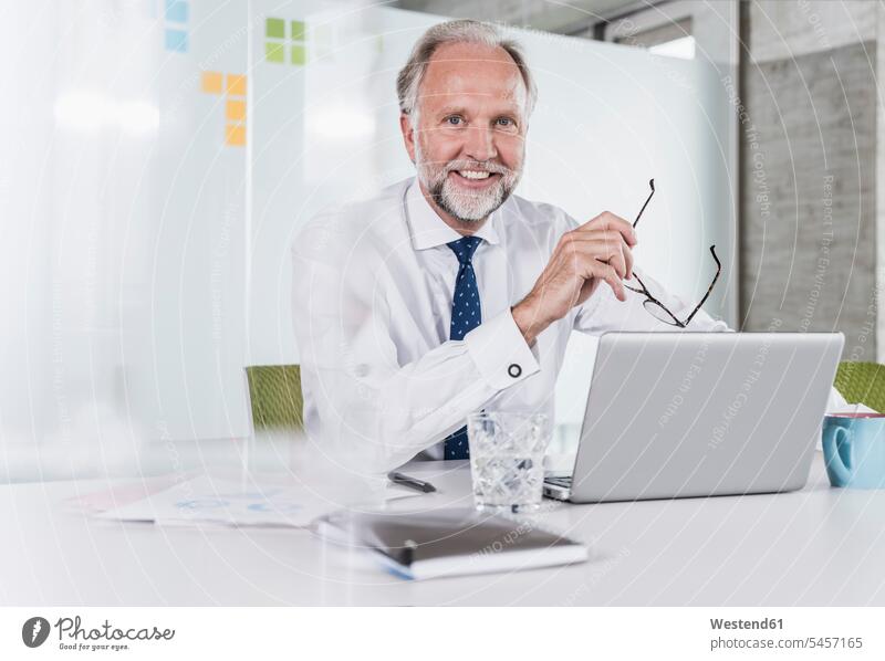 Porträt eines lächelnden reifen Geschäftsmannes, der mit Laptop am Schreibtisch im Büro sitzt Arbeitstisch Schreibtische Businessmann Businessmänner