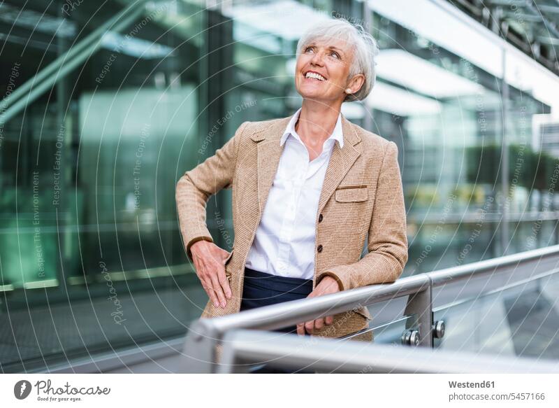 Lächelnde ältere Geschäftsfrau lehnt an einem Geländer in der Stadt und schaut nach oben Geschäftsfrauen Businesswomen Businessfrauen Businesswoman aufstützen