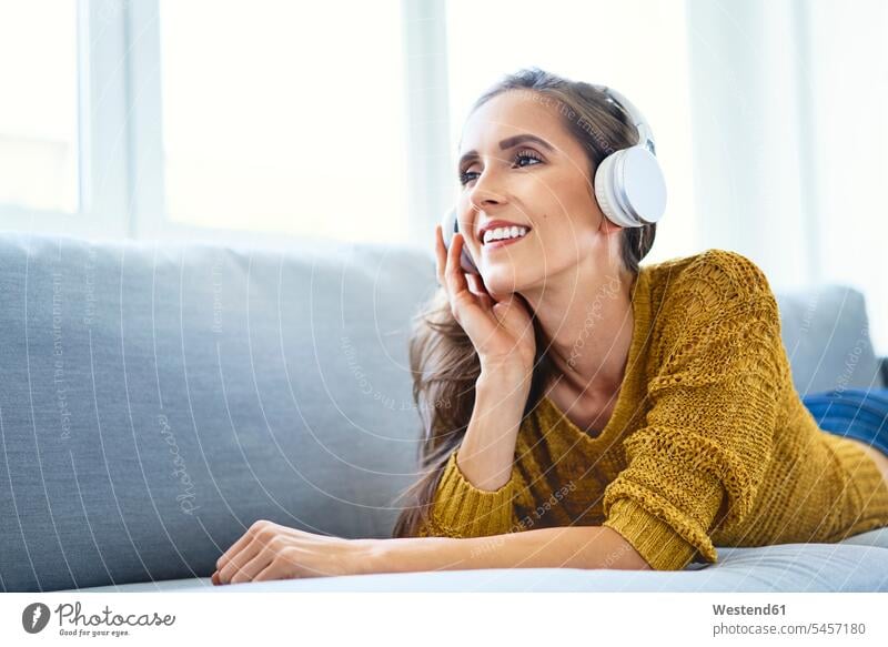 Glückliche junge Frau, die im Liegen auf dem Sofa mit Kopfhörern Musik hört Leute Menschen People Person Personen Alleinstehende Alleinstehender Singles