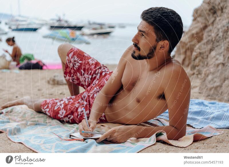 Junger Mann auf Handtuch am Strand liegend, Notizbuch, nachdenklich liegt nachdenken überlegen Tagebuch Tagebücher Tagebuecher Bart Bärte Männer männlich