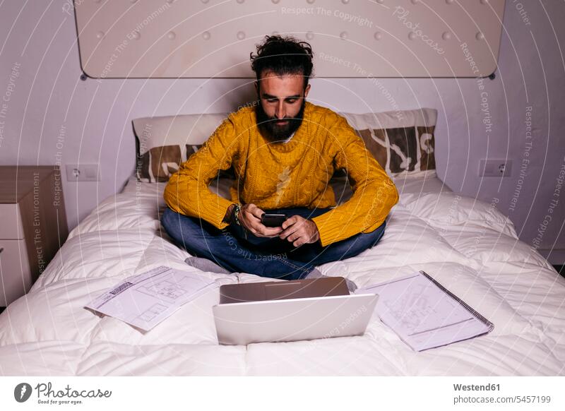Junger Mann arbeitet im Bett zu Hause mit Handy, Laptop und Dokumente Betten Papiere Unterlagen Notebook Laptops Notebooks Männer männlich arbeiten Arbeit