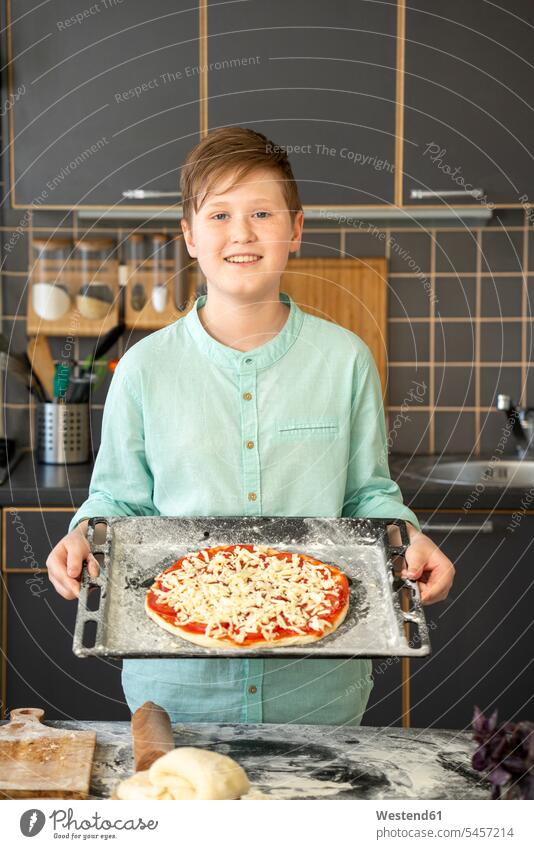 Porträt eines lächelnden Jungen, der ein Backblech mit roher hausgemachter Pizza hält Hemden freuen Glück glücklich sein glücklichsein stehend steht daheim