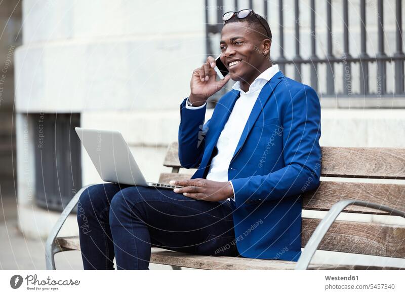 Junger Geschäftsmann in blauer Anzugjacke sitzt auf Bank und benutzt Smartphone Leute Menschen People Person Personen Afrikanisch Afrikanische Abstammung