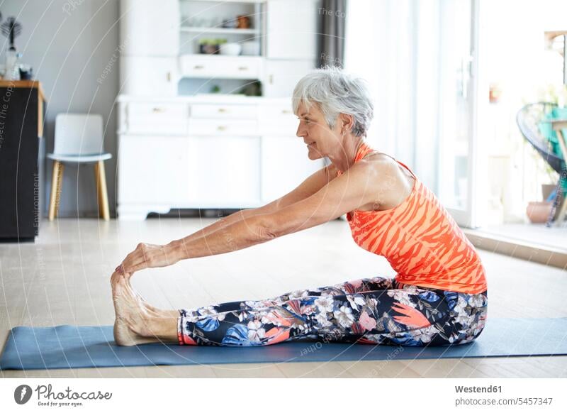 Passgenaue ältere Frau, die sich beim häuslichen Training Bein und Rücken streckt Farbaufnahme Farbe Farbfoto Farbphoto Innenaufnahme Innenaufnahmen innen