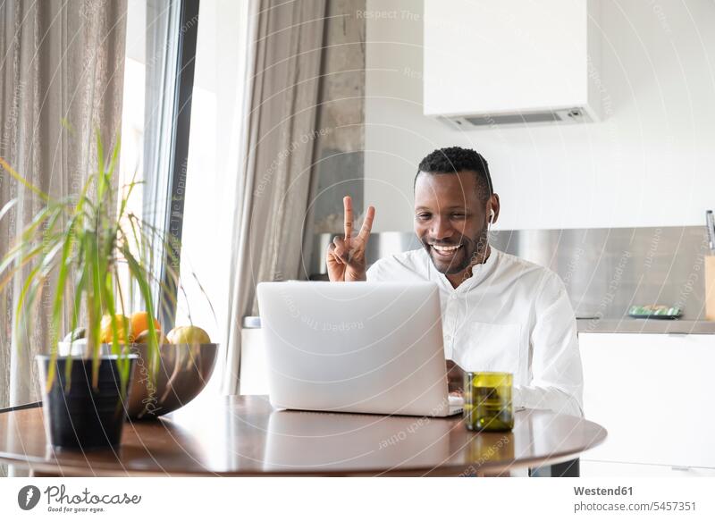 Porträt eines glücklichen Mannes, der während eines Video-Chats am Küchentisch sitzt und das Siegeszeichen zeigt Rechner Laptops Notebook Notebooks sitzend