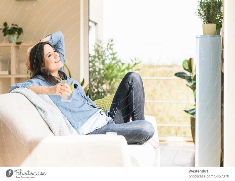 Glückliche Frau mit einem Glas aufgegossenem Wasser auf der Couch zu Hause sitzend Zuhause daheim weiblich Frauen sitzt Sofa Couches Liege Sofas Trinkgläser