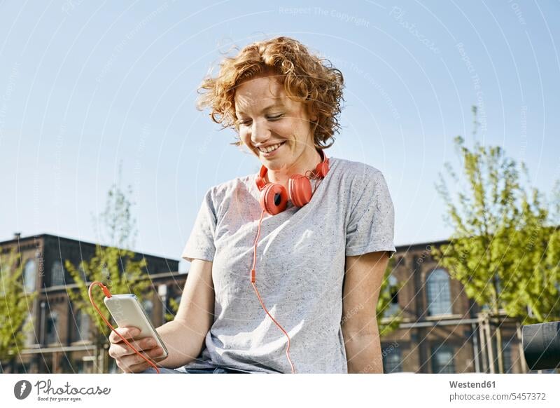 Lächelnde junge Frau mit Kopfhörern mit Smartphone in städtischer Umgebung Urban Urbanität Urbanitaet Kopfhoerer lächeln iPhone Smartphones weiblich Frauen