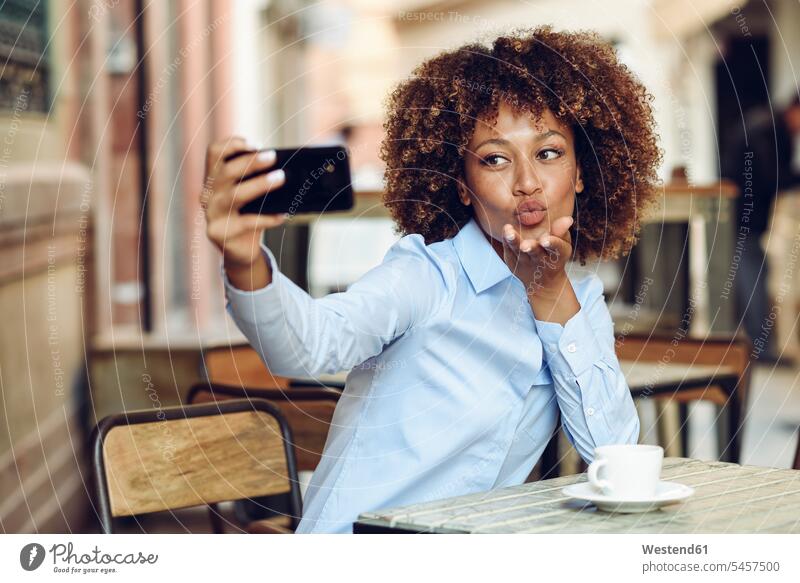 Frau mit Afrofrisur sitzt in einem Café im Freien und macht ein Selfie Selfies Afro-Look Afros Afrolook weiblich Frauen lächeln sitzen sitzend Cafe Kaffeehaus