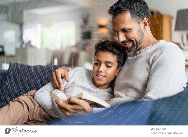 Vater liegt mit Sohn auf der Couch im Wohnzimmer und liest ein Buch Bücher Couches Liege Sofas knuddeln schmusen Lektüre sitzend sitzt entspannen relaxen