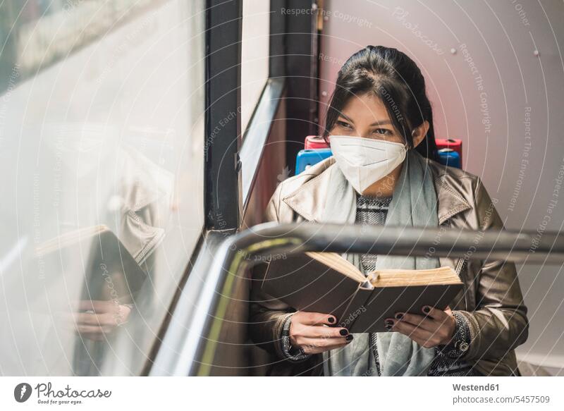Frau mit Schutzmaske, die durch ein Fenster schaut, während sie im Bus ein Buch liest Farbaufnahme Farbe Farbfoto Farbphoto Fahrzeuginnenraum