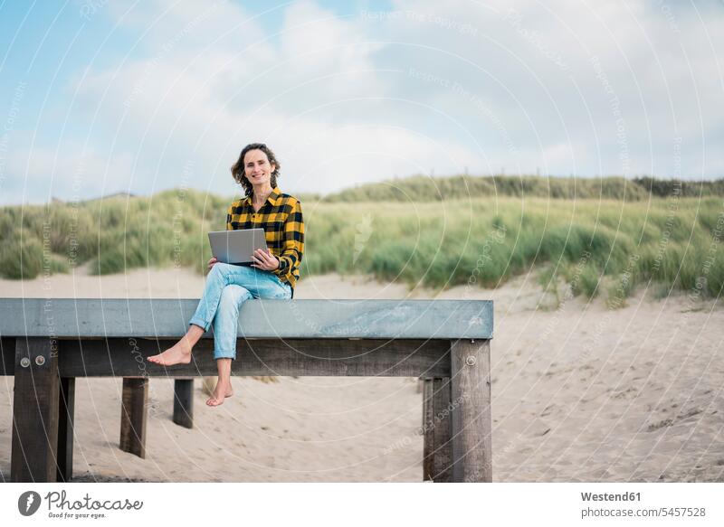 Reife Frau sitzt auf Promenade am Strand, mit Laptop Notebook Laptops Notebooks benutzen benützen Holzsteg Holzstege Steg Stege Beach Straende Strände Beaches
