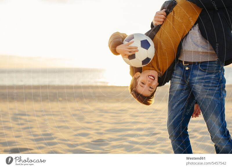 Vater hält Sohn mit auf dem Kopf stehendem Fussball bei Sonnenuntergang am Strand Söhne halten Sonnenuntergänge Fußball Fußbälle Beach Straende Strände Beaches