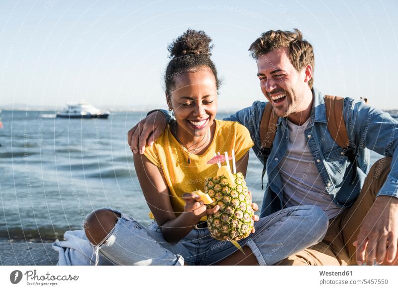Glückliches junges Paar sitzt am Pier am Wasser mit einer Ananas, Lissabon, Portugal Leute Menschen People Person Personen Afrikanisch Afrikanische Abstammung