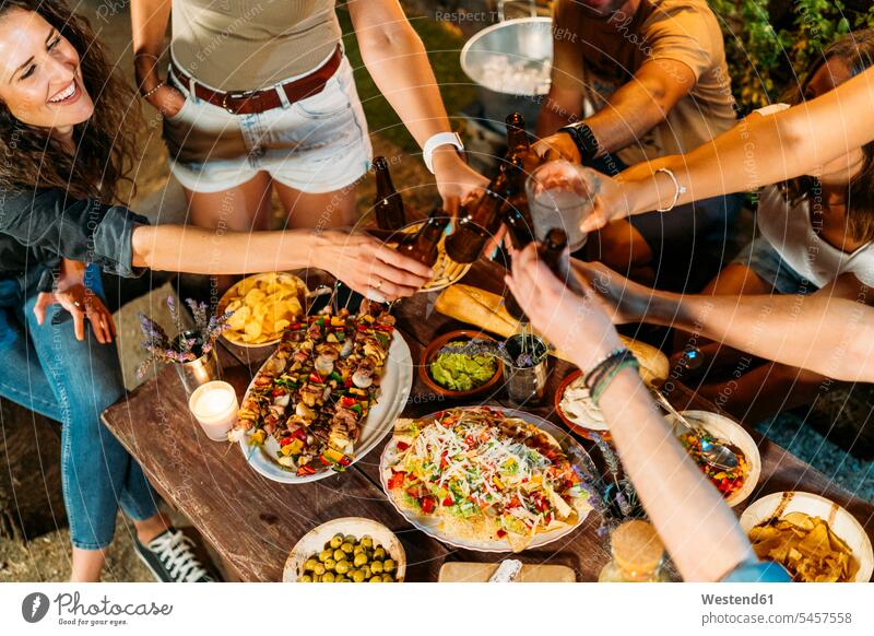 Glückliche Freunde stoßen bei einem Abendessen im Freien mit Bierflaschen an Kameradschaft Flaschen Tische Holztische entspannen relaxen abends nachts