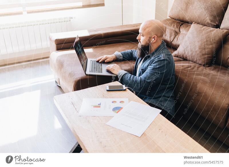 Geschäftsmann mit Diagrammen, der auf seinem Smartphone Musik hört, während er zu Hause einen Laptop benutzt Farbaufnahme Farbe Farbfoto Farbphoto